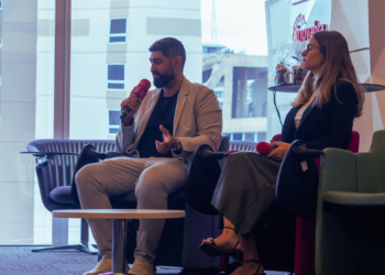 Adriana Tedeschi e José Parelho sentados em poltronas, segurando microfones e participando de um painel em um ambiente corporativo, com vista para prédios ao fundo.
