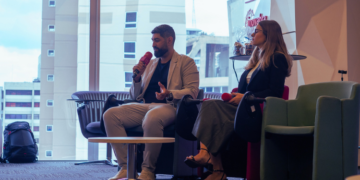 Adriana Tedeschi e José Parelho sentados em poltronas, segurando microfones e participando de um painel em um ambiente corporativo, com vista para prédios ao fundo.