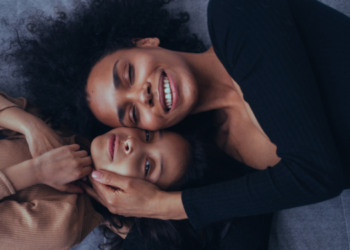 Mãe negra deitada ao lado de sua filha, ambas sorrindo e abraçadas carinhosamente.