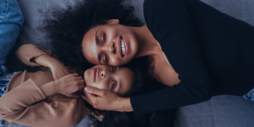 Mãe negra deitada ao lado de sua filha, ambas sorrindo e abraçadas carinhosamente.