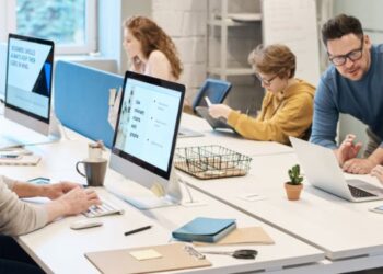 Pessoas trabalhando em equipe em um escritório moderno, com computadores, tablets e materiais de trabalho sobre a mesa.