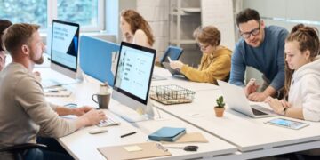 Pessoas trabalhando em equipe em um escritório moderno, com computadores, tablets e materiais de trabalho sobre a mesa.